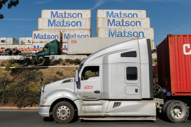 A gray and white commercial truck drives down a street. Another green and white commercial truck can be seen up a hill in the distance alongside several cargo containers labeled 