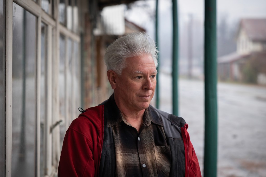 A person with neatly styled silver hair stands under a covered walkway, looking to the side with a calm expression. They wear a red and black jacket over a plaid shirt. The background shows a rainy, quiet street with buildings and telephone poles fading into the distance. The atmosphere feels still and contemplative.