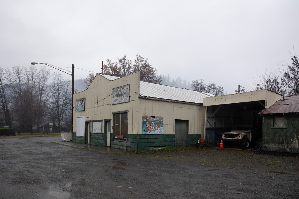 A foggy, overcast day in a rural town shows a weathered cream and green building labeled โGuide Service.โ The small structure has boarded-up windows, a faded sign with a phone number, and a hand-painted โWhite Water Toursโ mural on the side. To the right is an open garage-like shelter with an old, rusted vehicle and an orange traffic cone. Bare trees and mist-covered hills fill the background.