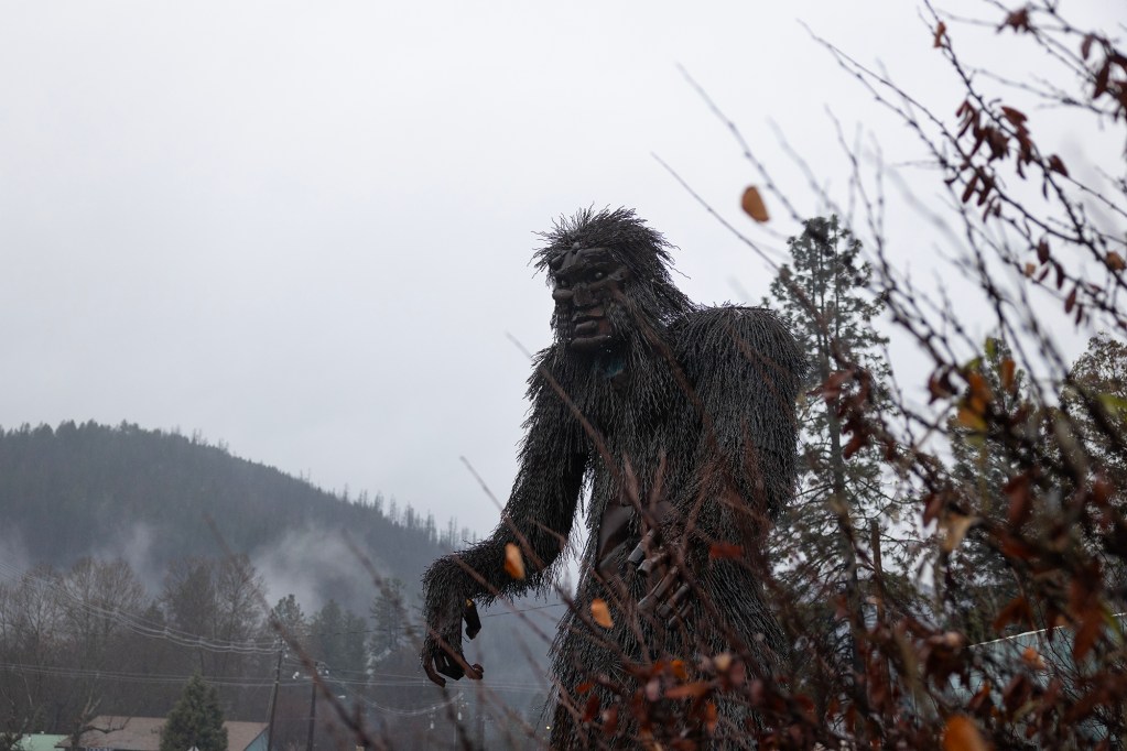 A towering sculpture of a Bigfoot-like creature made from dark, textured metal stands among bare branches in a misty, forested area. The figure has exaggerated features and a shaggy appearance, with one arm extended forward. Pine trees, power lines, and rooftops are visible in the background beneath an overcast sky, adding to the eerie atmosphere.