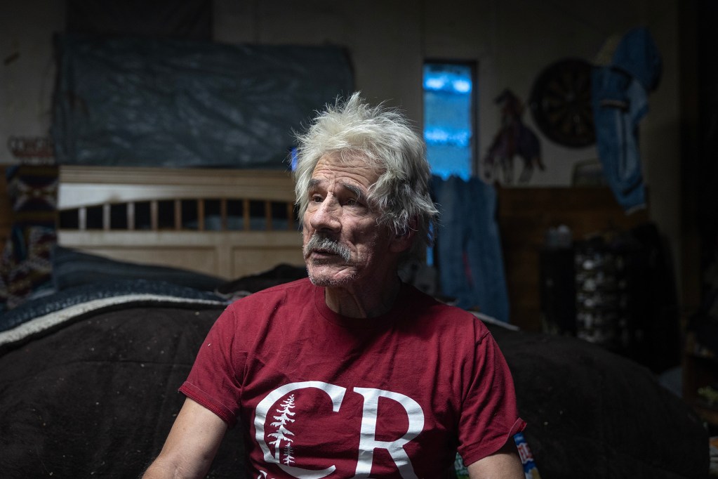 An older person with white hair and a mustache sits indoors on the edge of a bed, wearing a red t-shirt with a white "CR" logo. The room is dimly lit, with personal items and decorations visible in the background, including a dartboard and a painting of a horse and rider.