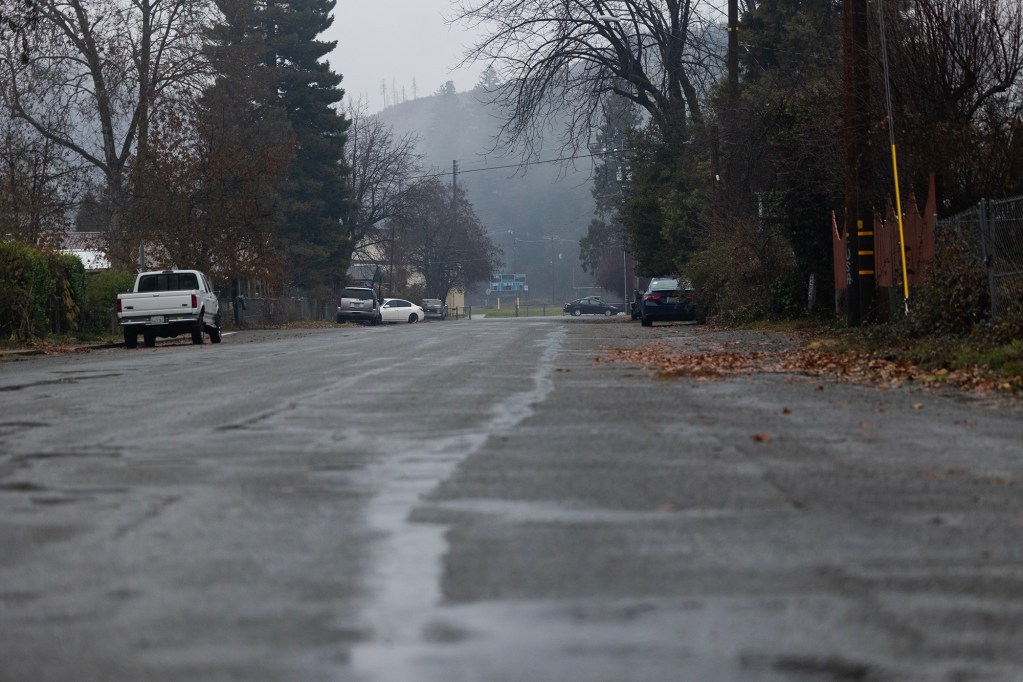 A wet, empty street in a small town on a foggy day, with a few parked cars along the sides and leafless trees lining the road. Low hills and more trees are visible in the misty background.