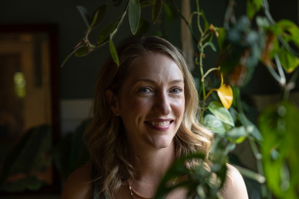 A person with long wavy blond hair smiles while standing indoors, framed by green leafy houseplants. Soft natural light highlights their face, while the background remains dim, creating a warm and intimate portrait.