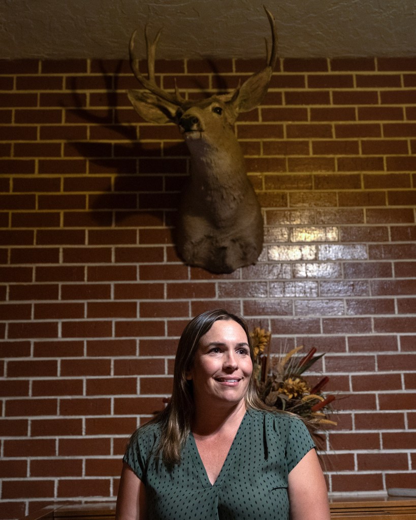A woman in a green blouse sits indoors against a red brick wall, smiling slightly while looking off-camera. Mounted above her on the wall is a taxidermy deer head with antlers, casting a shadow.