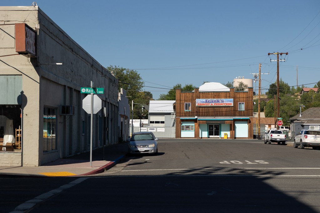 A quiet downtown street in Alturas, California, with a Main Street sign in the foreground and a brown building with a sign reading "Frank’s Carpet & Furniture" in the distance. A few parked cars and utility poles line the street under clear blue skies.
