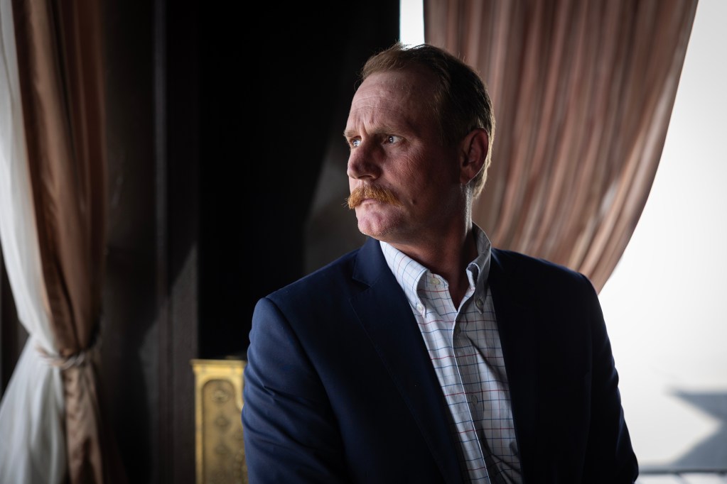 A person with light skin, short hair, and a reddish mustache wearing a navy blue blazer over a checkered dress shirt stands indoors near a window with draped curtains, gazing out with a serious expression.