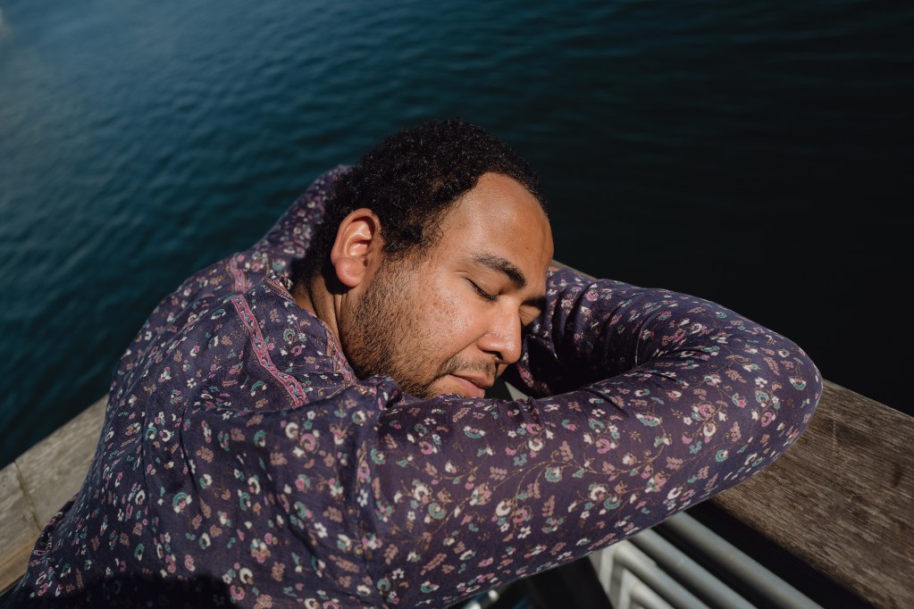 A person closes their eyes and lays their head against a wooden rail overlooking a waterfront.