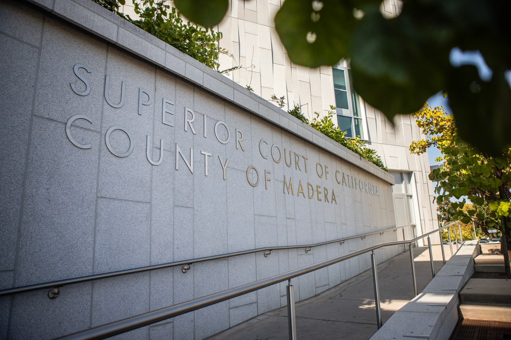 A long concrete wall outside a courthouse displays the raised lettering “Superior Court of California, County of Madera,” with a wheelchair-accessible ramp and handrail running alongside it, partially framed by leafy trees.