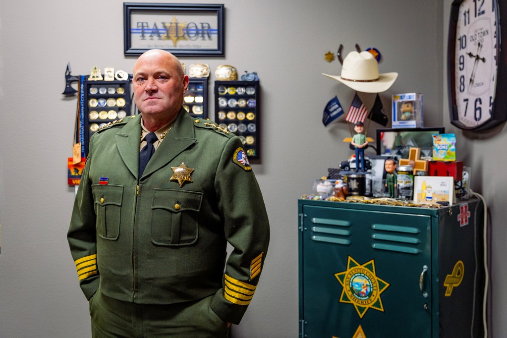 A uniformed law enforcement officer stands with hands in pockets inside an office decorated with badges, patches, memorabilia, and a sheriff’s emblem. A cabinet with a sheriff’s star logo, small figurines, flags, and a cowboy hat sits behind them alongside framed badge displays on the wall.