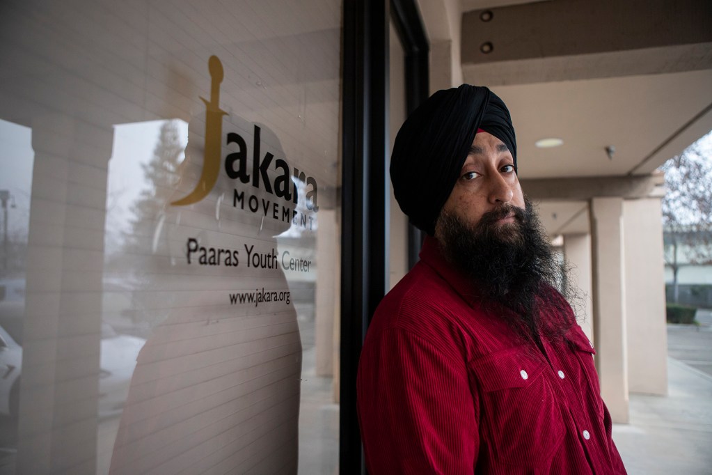 A person wearing a black turban and red jacket stands beside a glass door displaying the Jakara Movement and Paaras Youth Center logo, looking toward the camera along a covered walkway outside the building.