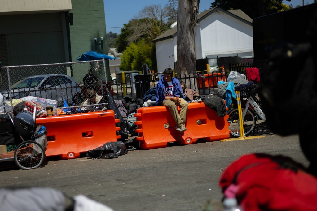 An unhoused person sits on top of an orange safety barrier surrounded by various posessions alongside a street.