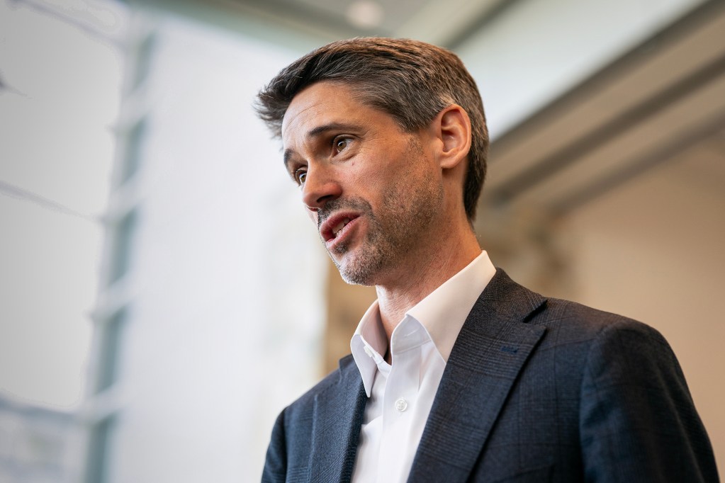 A person in a dark blazer and white dress shirt speaks while turned slightly to the side. They're at an indoor conference event space.