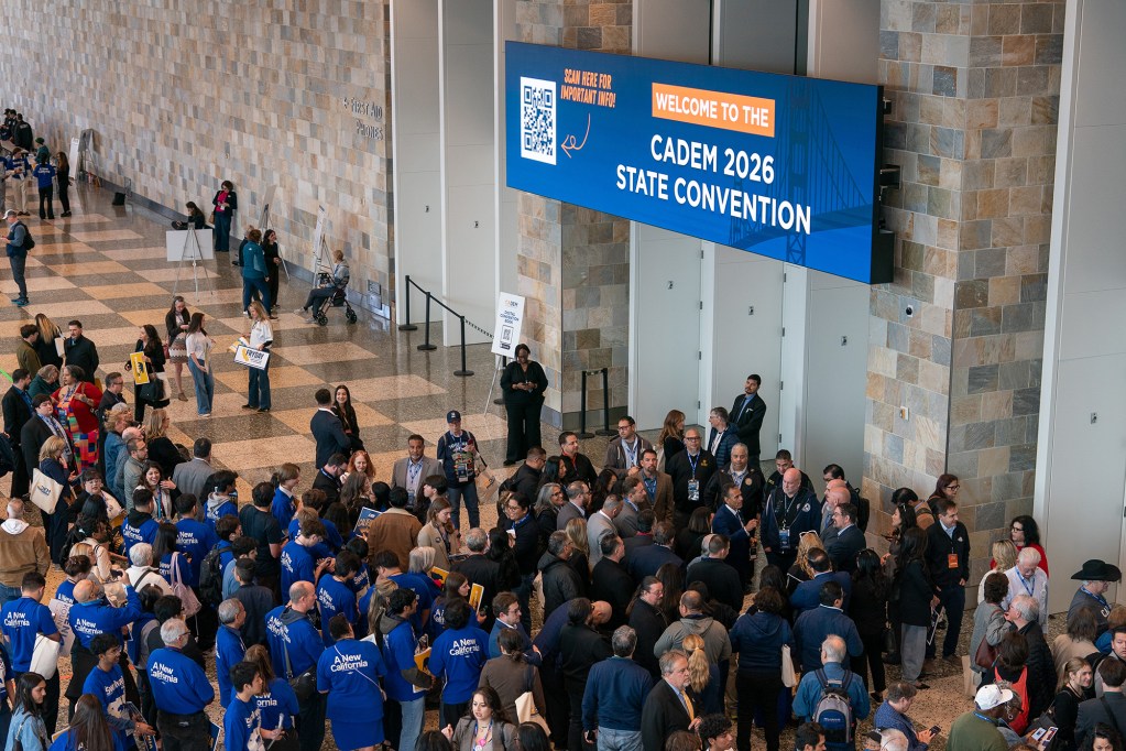 A large crowd gathers inside a spacious convention hall beneath a blue sign that reads “Welcome to the CADEM 2026 State Convention.” Attendees cluster in groups across the tiled floor, many wearing badges and blue shirts, talking, waiting, or looking toward a central area. The setting has tall stone-patterned walls, high ceilings, and wide doorways, giving the scene a formal conference atmosphere.