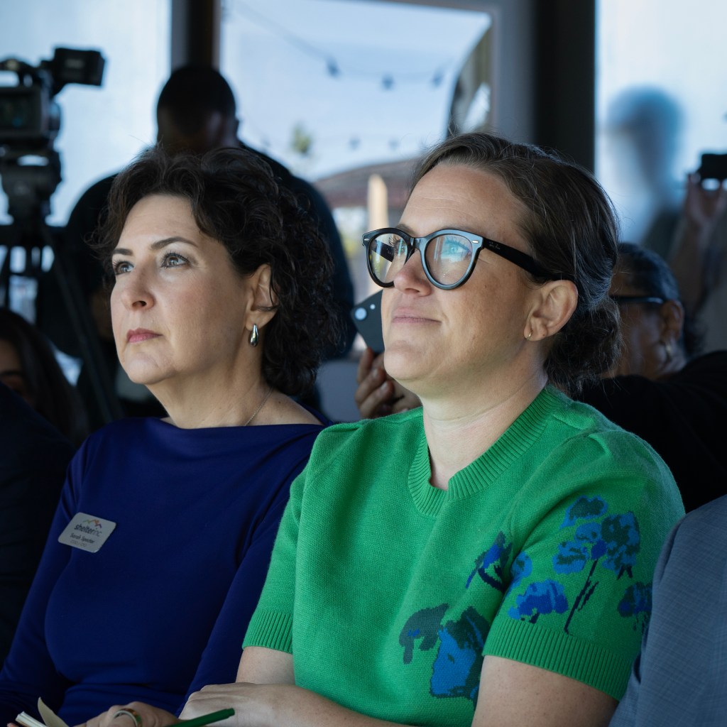Two people sit side by side listening attentively at an indoor event, with cameras and other attendees visible in the background.