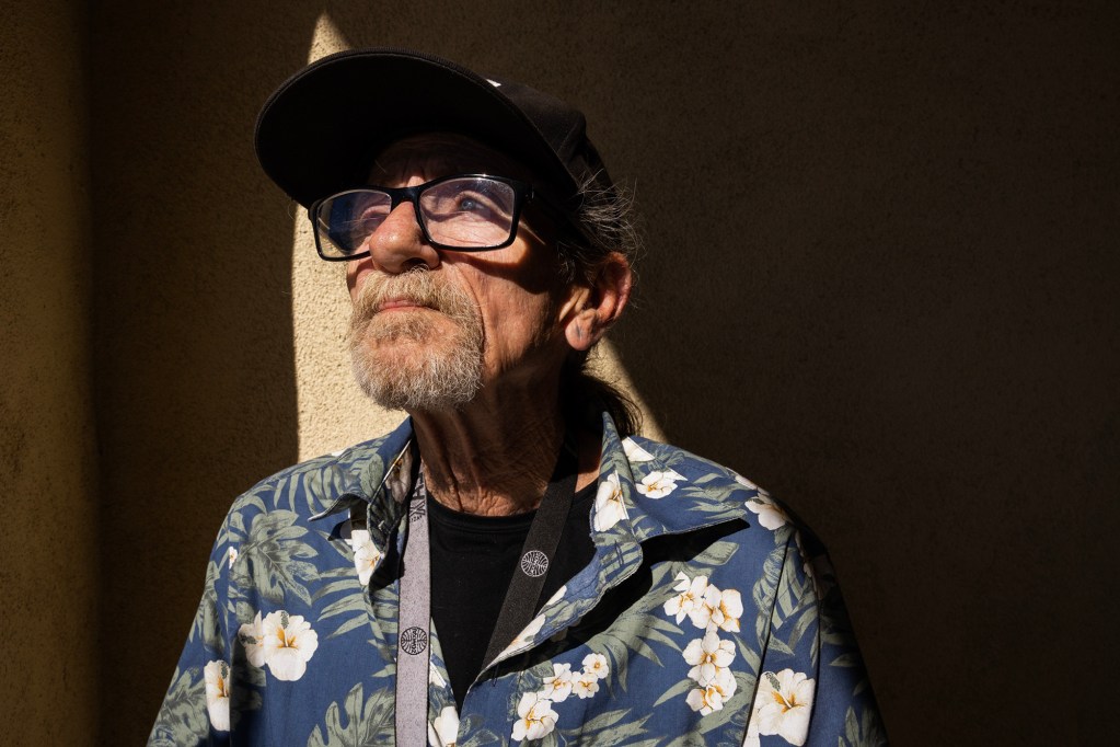 An older adult wearing glasses, a baseball cap, and a floral shirt stands in strong sunlight, looking upward against a textured wall.