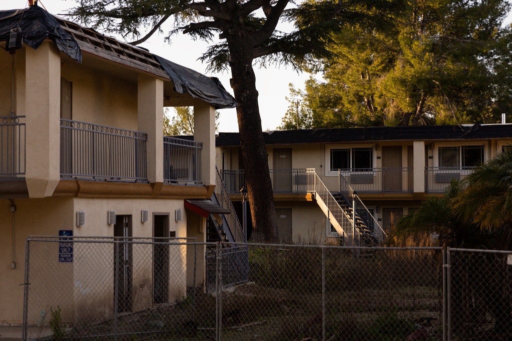An exterior view of a two-story motel-style building behind a chain-link fence, with damaged roofing partially covered by black tarp, exterior staircases and walkways, and trees casting warm late-day light across the scene.