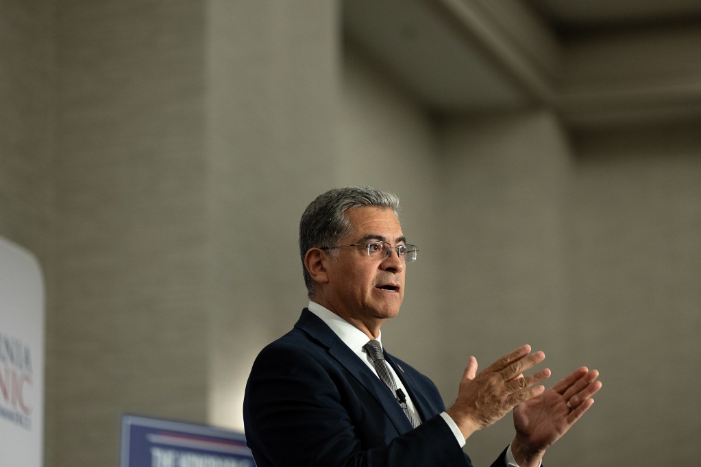 A side view of a person, with short gray hair and wearing a blue suit, gestures with both their hands as they stand on a small stage set against a beige wall inside a large room.